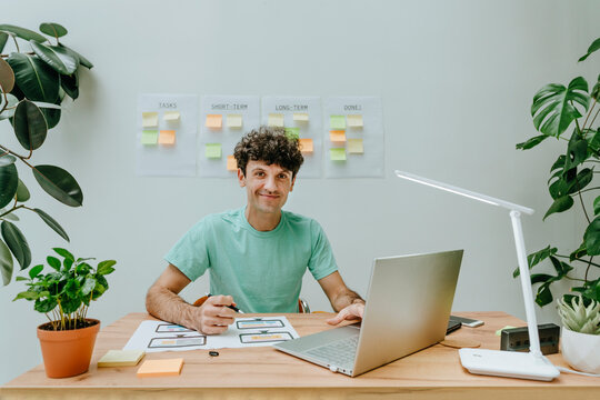 Smiling graphic designer sitting with laptop and printout at desk in office