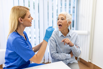 Senior patient opening her mouth for the doctor to look in her throat. Female doctor examining sore throat of patient in clinic. Otolaryngologist examines sore throat of patient. - Powered by Adobe