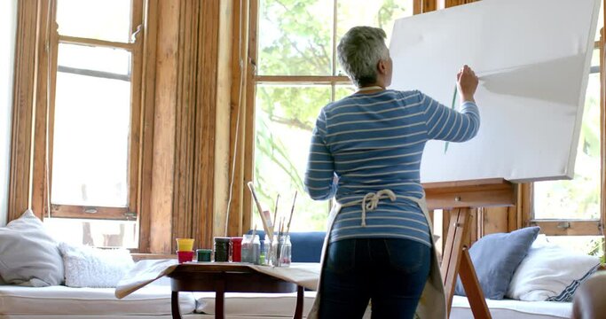 Senior biracial woman wearing apron and painting on big canvas at home, slow motion