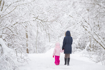 Mother and daughter walking on white fresh snow covered road through tree branches at park after...