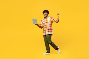 Full body young excited IT Indian man he wears shirt casual clothes hold use work on laptop pc computer do winner gesture isolated on plain yellow color background studio portrait. Lifestyle concept.