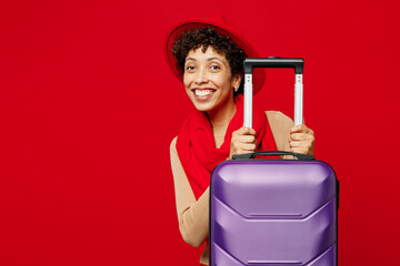 Traveler woman wear sweater hat scarf hold purple suitcase bag looking camera isolated on plain red background. Tourist travel abroad in free spare time rest getaway. Air flight trip journey concept.