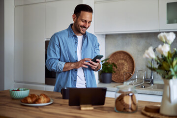 A smiling bearded adult man looks for a recipe on a phone while standing in a kitchen.
