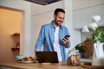 A cheerful adult man scrolls through social media on a phone while standing in a kitchen.