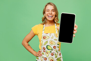 Young smiling housewife housekeeper chef cook baker woman wear apron yellow t-shirt hold use blank screen mobile cell phone blink eye isolated on plain pastel green background. Cooking food concept.