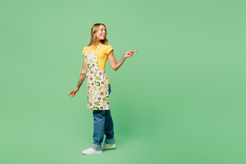 Full body side view young housewife housekeeper chef cook baker woman wear apron yellow t-shirt point index finger aside on area mock up isolated on plain green background studio Cooking food concept