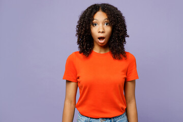 Little shocked panicked sad kid teen girl of African American ethnicity she wear orange t-shirt look camera with opened mouth isolated on plain pastel purple background. Childhood lifestyle concept.