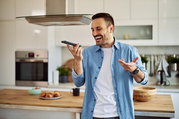 A joyful adult man laughing and joking with his friend over a phone call in a kitchen.