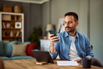 A confident male freelancer scrolling through social media on his phone while sitting in front of a tablet for his work.