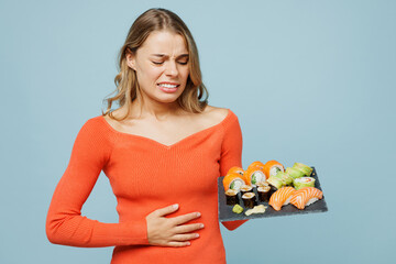 Young sad sick ill woman he wear orange casual clothes puts hand on tummy belly holding eat raw fresh sushi roll served on black plate Japanese food isolated on plain blue background studio portrait.