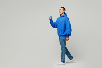 Full body young middle eastern man wear blue hoody casual clothes hold takeaway delivery craft paper brown cup coffee to go isolated on plain solid white background studio portrait. Lifestyle concept.