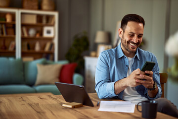 A cheerful man texts his friends on his phone while taking a break from his work on tablet.