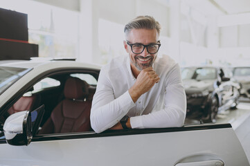 Adult man customer male buyer client wearing shirt glasses open lean on door prop up chin choose auto want buy new automobile in car showroom vehicle salon dealership store motor show. Sales concept. - obrazy, fototapety, plakaty