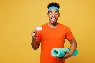 Young amazed fitness trainer instructor sporty man sportsman wear orange t-shirt hold caremat credit bank card spend time in home gym isolated on plain yellow background Workout sport fit abs concept
