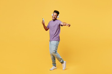 Full body fun young man of African American ethnicity he wears purple t-shirt casual clothes use mobile cell phone listen to music in headphones isolated on plain yellow background. Lifestyle concept.