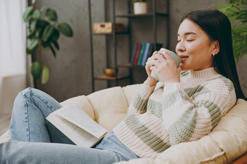 Sideways young calm smart fun woman wear casual clothes sits on armchair read book novel drink tea stay at home hotel flat rest relax spend free spare time in grey living room indoor. Lounge concept. - obrazy, fototapety, plakaty
