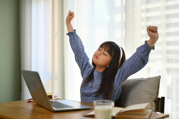 Tired schoolgirl stretching in front of laptop while studying remotely at home. Online education...