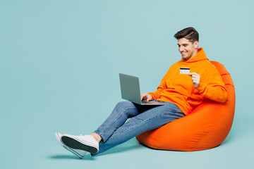 Full body smiling young man wears orange hoody casual clothes sit in bag chair using mobile cell...