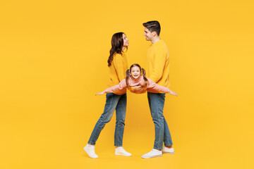 Full body young smiling fun parents mom dad with child kid girl 7-8 years old wear pink sweater casual clothes hold daughter pov she is flying isolated on plain yellow background. Family day concept.