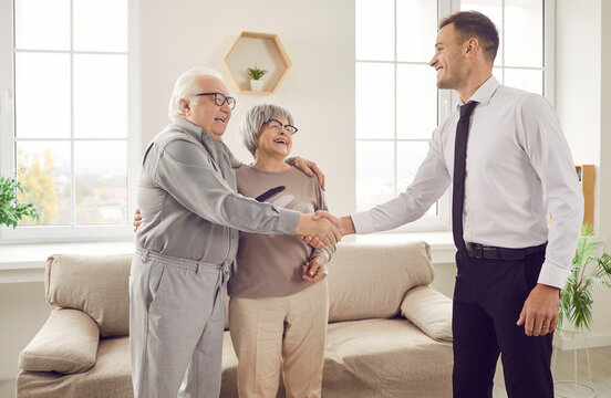 Portrait of happy cheerful senior couple standing in the living room at home shaking hands and thanking male financial advisor reaching agreement in health insurance. Investment in retirement concept - Powered by Adobe