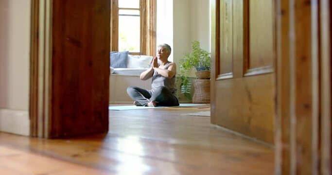 Focused senior biracial woman meditating on yoga mat at home