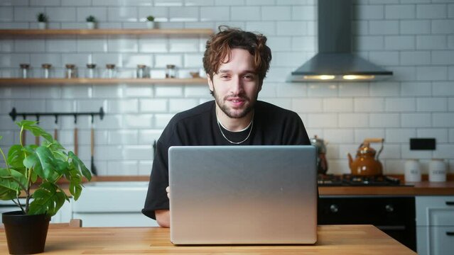 Young curly hair vlogger man sitting in kitchen with laptop computer, catch the gift box in the air, advertising product , talking to camera, recording video
