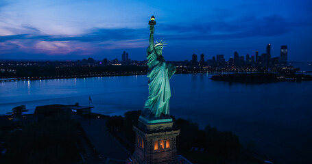 Aerial Helicopter Cinematic View of the Statue of Liberty with Manhattan Skyline Cityscape in the Evening. Panoramic View of New York City Skyscrapers and Jersey City Buildings at Sunset