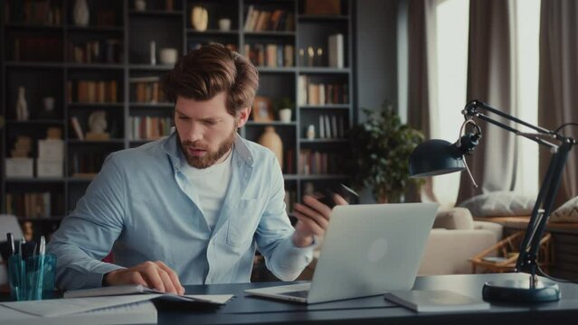 young man with beard in blue shirt sits at table in home office, multitasking with laptop, mobile phone, notebook, be in a hurry, stays focused under the pressure work deadlines. speed up
