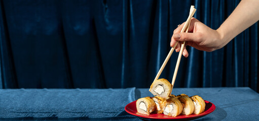 Female hand with chopsticks holding sushi roll on blue velvet background. Roll with salmon on red plate. Modern, stylish Japanese cuisine. space for text