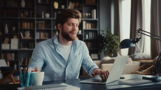 speed up portrait of young man with beard in blue shirt sits at table in home office, typing on keyboard non stop, smiling and staying focused