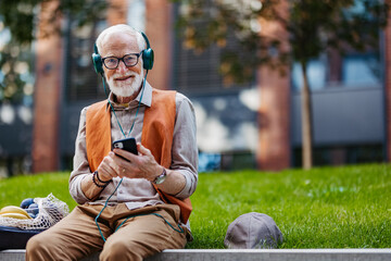 Stylish senior man sitting in the city park, listening to music via headphones. Concept of old man young at heart.