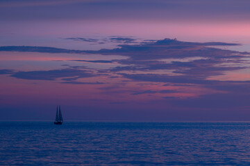A single sailboat on the ocean, against a tropical, purple sunset sky, at dusk