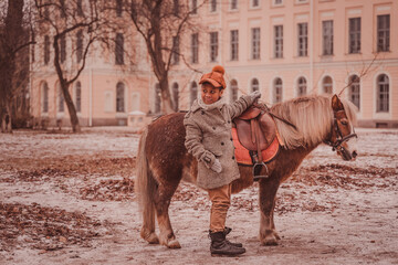 a boy stands next to a pony, holding the pommel of the saddle, looking back suspiciously