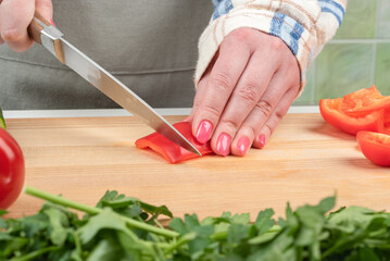 Female hands with manicure cut a piece of sweet pepper on a wooden cutting board.