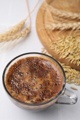 Cup of barley coffee, grains and spikes on white table, closeup