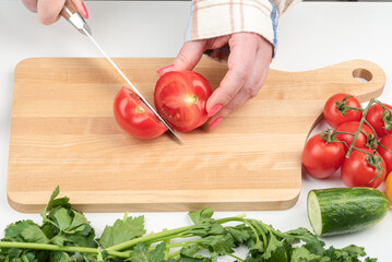 A young woman with a fashionable manicure uses a knife to cut a tomato on a wooden cutting board.