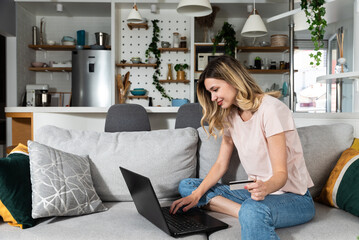 Young woman sitting on the sofa using laptop computer and credit card for online shopping and ordering groceries and food for home so she dont have to go out to buy food.