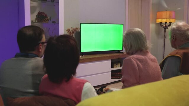 Family watching TV. Green screen. Two couples, an elderly and a middle-aged couple, are sitting on the couch at home. In front of them is a green screen TV. Horizontal panning. 