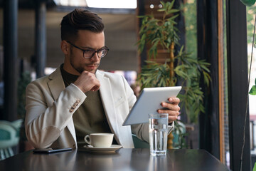 Serious businessman wearing suit and eyewear using digital tablet sitting at table in a Cafe,...