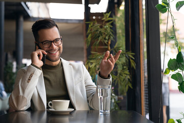Happy wealthy successful young business man talking on the phone in Cafe. Smiling professional businessman executive entrepreneur wearing suit making corporate call on cellphone. - obrazy, fototapety, plakaty