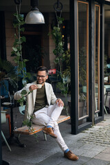 Happy wealthy successful young business man enjoying morning coffee while sitting on a swing in...