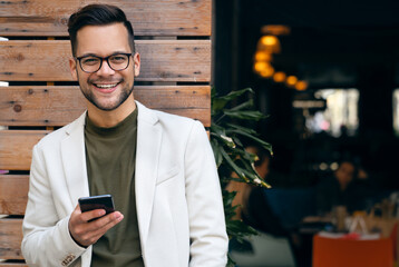 Portrait of young man professional CEO looking at camera and using smartphone while standing in front of restaurant. - obrazy, fototapety, plakaty