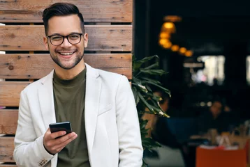 Tuinposter Portrait of young man professional CEO looking at camera and using smartphone while standing in front of restaurant. © Dorde