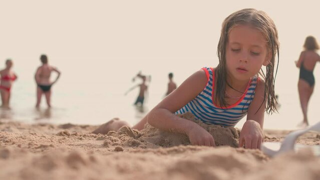 Slow motion shot of cute little girl dressed in stripped outfit playing with sand on beach near waving sea at sunset during vacation