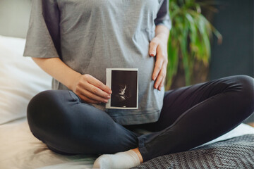 Cropped shot of pregnant woman holding pregnancy ultrasound while sitting in lotus pose on bed at...