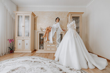 Morning of the bride before the wedding. A beautiful young woman with a veil and a beautiful hairstyle in a white robe near her wedding dress on a mannequin. Professional makeup. Last preparations.