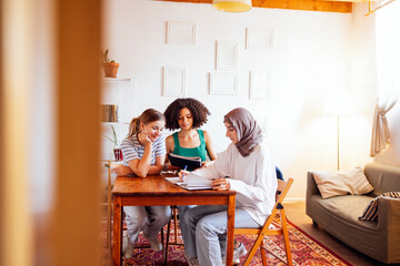 Three cute teenage girls do their homework. Caucasian, African American and a Muslim female teens sit at a table with books and notebooks. White wall and plant pots on background. Copy space.