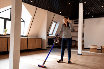 Happy brunette woman listening music with headphones and dancing while vacuum cleaning floor in a cozy room