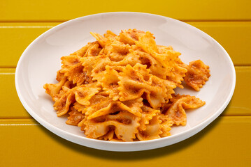 Farfalle pasta with tomato and chili and garlic sauce and olive oil in white plate isolated on yellow wooden table with clipping path included