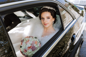Portrait of an incredibly beautiful bride in the window of an expensive car. The bride with a bouquet of flowers looks into the camera lens.